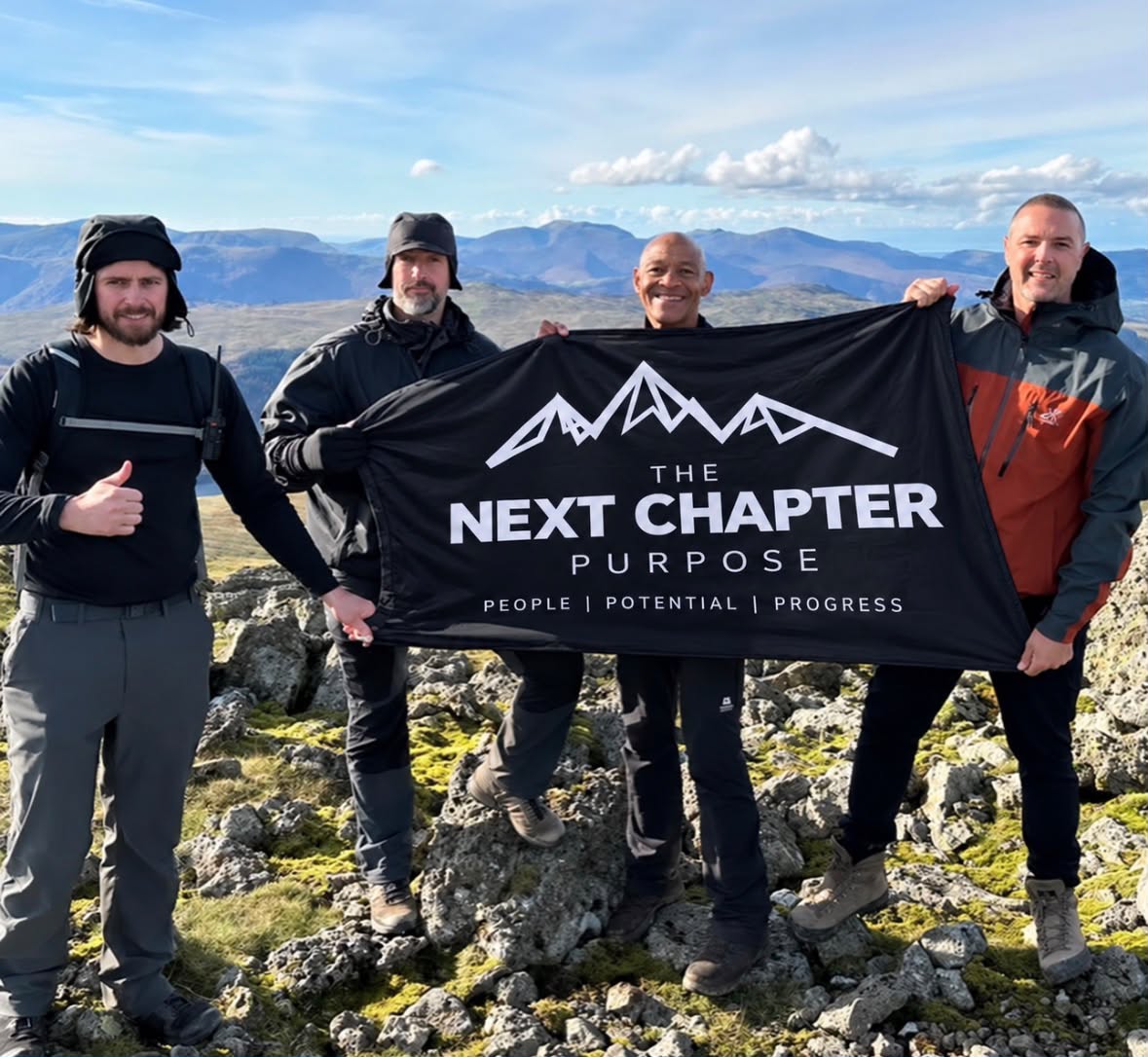 Four people on a mountain holding The Next Chapter PURPOSE flag
